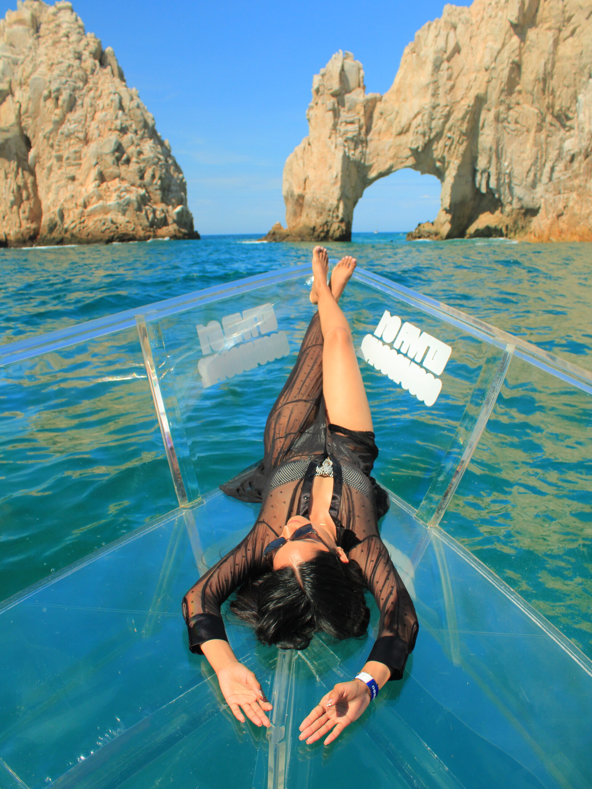 Model Karina Araujo posing on a transparent boat in front of the Cabo San Lucas Arch